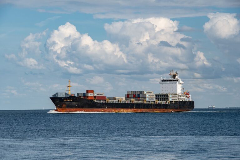 A large cargo ship sailing on the ocean with clear blue skies and white clouds above.