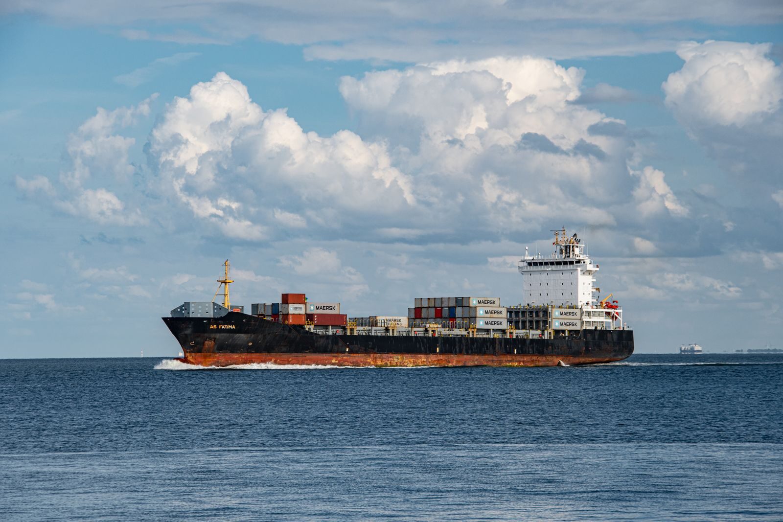 A large cargo ship sailing on the ocean with clear blue skies and white clouds above.